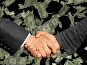 Two businessmen engage in a handshake above a substantial pile of money, representing a lucrative business deal