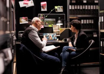 Two people seated at a desk in an office, surrounded by files and paperwork